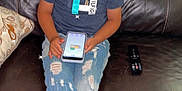 Bellamy is registered to the contest to win money with this photo: child, laughing, smiling, blue_shirt, ripped_jeans, sneakers, smartphone, couch, remote_control, blanket, indoor, happy, jewelry, sitting, brown_leather, floor, casual_clothing, home, portrait, joyful