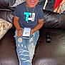 child, laughing, smiling, blue_shirt, ripped_jeans, sneakers, smartphone, couch, remote_control, blanket, indoor, happy, jewelry, sitting, brown_leather, floor, casual_clothing, home, portrait, joyful