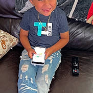 Bellamy is registered to the contest to win money with this photo: child, boy, smiling, couch, smartphone, jeans, tshirt, necklace, remote_control, blanket, indoor, casual, happy, seated, ripped_jeans, footwear, home, playing, technology, portrait