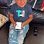 child, boy, smiling, couch, smartphone, jeans, tshirt, necklace, remote_control, blanket, indoor, casual, happy, seated, ripped_jeans, footwear, home, playing, technology, portrait
