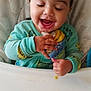 baby_food, child, closed_eyes, cute, face, feeding, food_pouch, green_clothing, hand, happy, high_chair, indoor, infant, messy, person, seat, smiling, snack, table, toddler