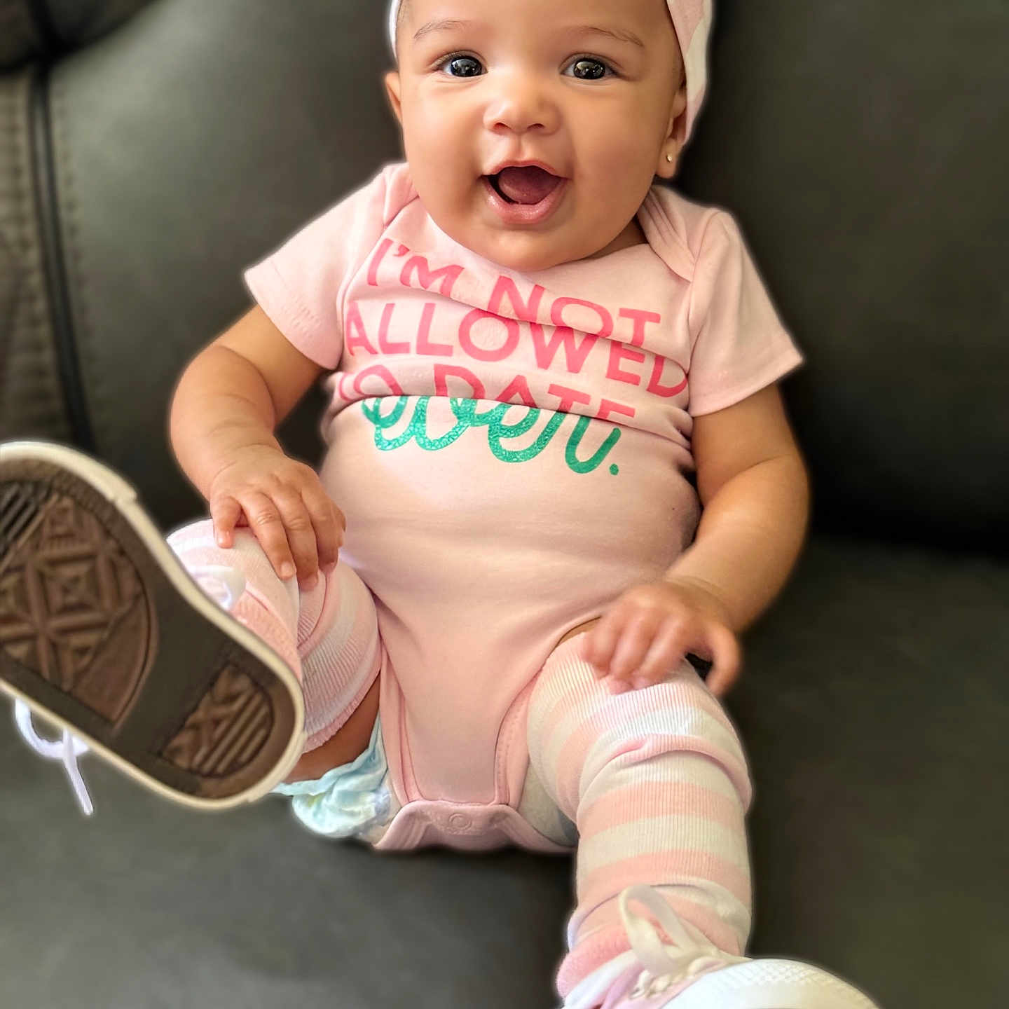 Navy is registered to the contest to win money with this photo: baby, child, clothing, couch, cute, face, footwear, happy, headwear, indoor, infant, onesie, pink_bow, playful, portrait, sitting, smiling, sneakers, striped_leg_warmers, toddler