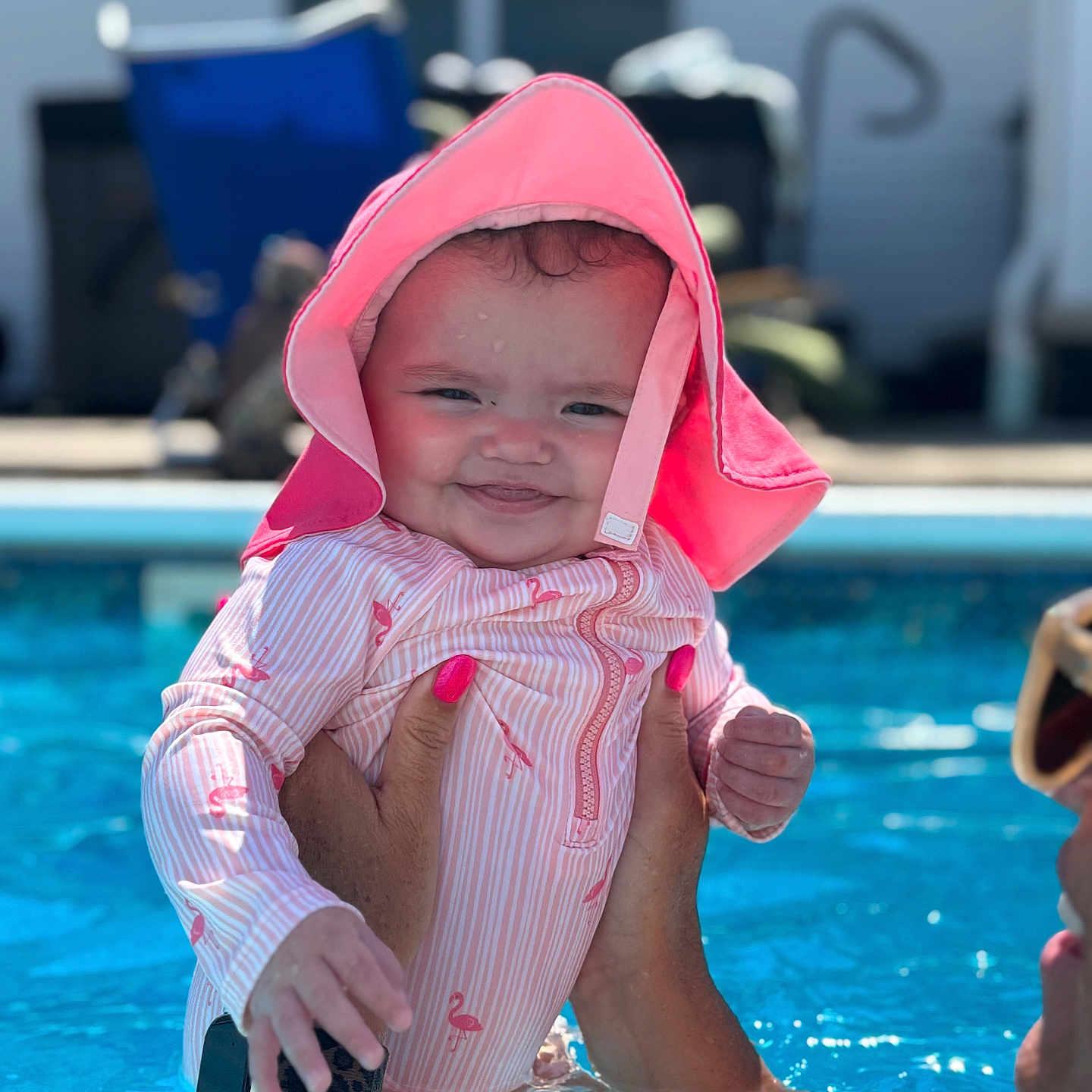 Zuri Lynn is registered to the contest to win money with this photo: baby, child, closeup, face, flamingo_pattern, fun, hand, happy, holding, nail_polish, outdoor, person, pink_hat, playful, smile, summer, sunny, swimming_pool, swimsuit, water