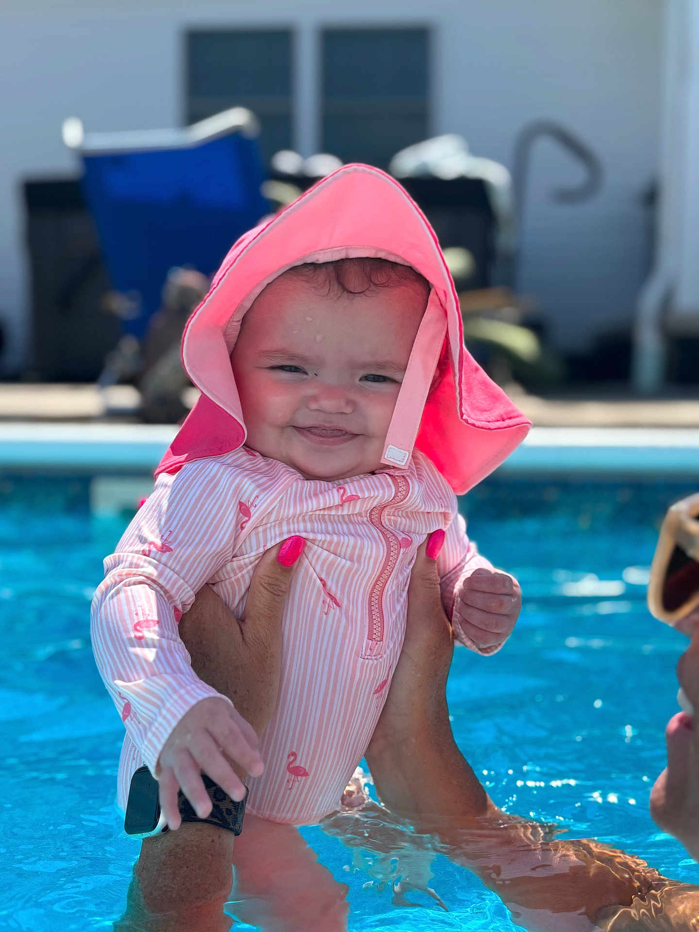 Zuri Lynn is registered to the contest to win money with this photo: baby, child, swimming_pool, water, pink_hat, swimsuit, flamingo_pattern, person, hand, nail_polish, smile, sunny, outdoor, summer, fun, playful, face, holding, closeup, happy