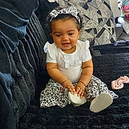 Akeilah is registered to the contest to win money with this photo: baby, child, smiling, sitting, white_clothing, floral_pattern, headband, couch, indoor, living_room, blanket, window, toy, person, happy, curly_hair, footwear, black_couch, cute, home