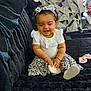 baby, child, smiling, sitting, white_clothing, floral_pattern, headband, couch, indoor, living_room, blanket, window, toy, person, happy, curly_hair, footwear, black_couch, cute, home