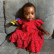 Star is registered to the contest to win money with this photo: toddler, child, red_dress, hearts_pattern, gray_couch, toy, grape_toy, sitting, indoor, curly_hair, person, young_child, casual, furniture, small_hand, barefoot, portrait, cute, comfortable, expression