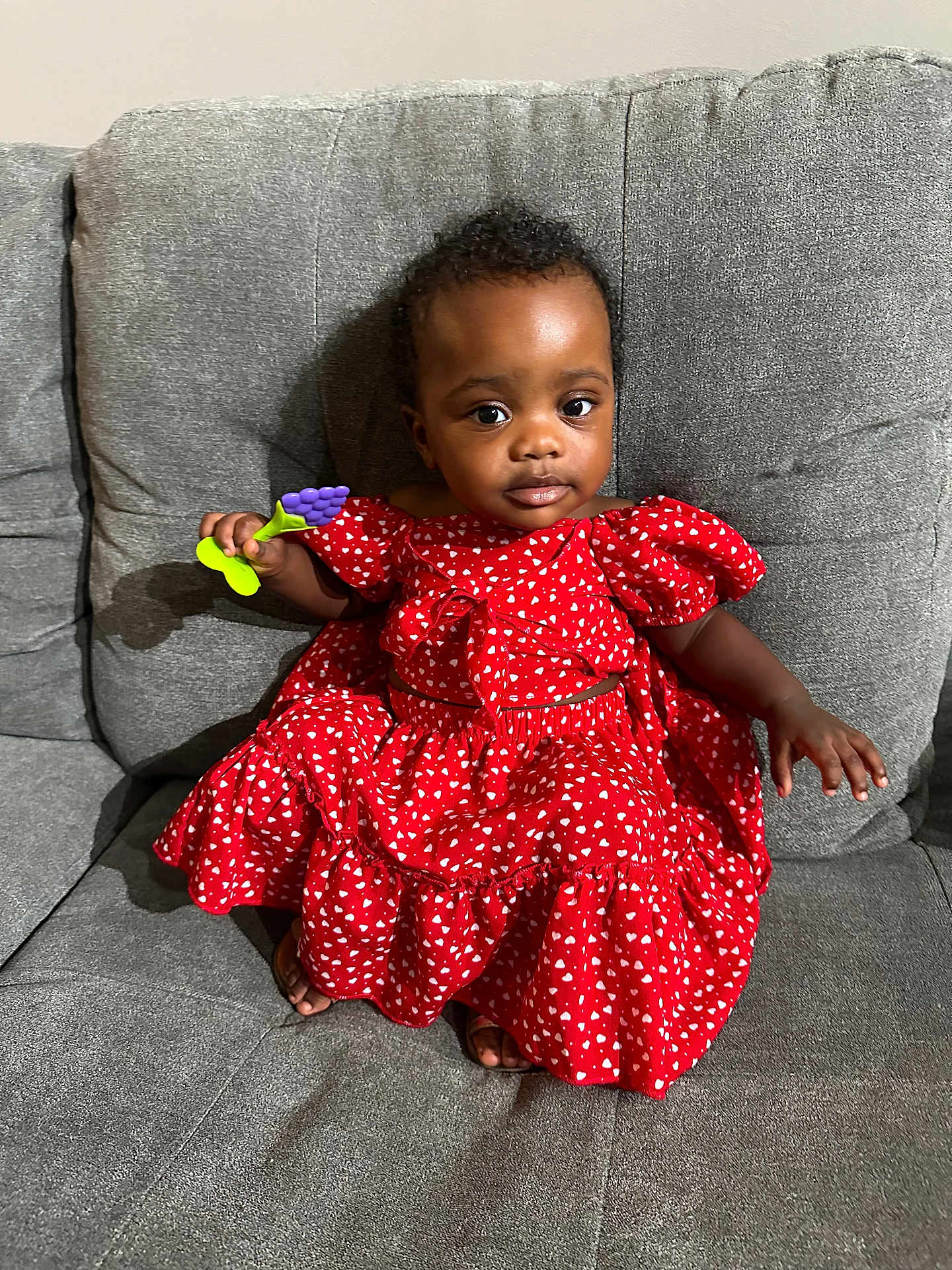 Star is registered to the contest to win money with this photo: toddler, child, red_dress, hearts_pattern, gray_couch, toy, grape_toy, sitting, indoor, curly_hair, person, young_child, casual, furniture, small_hand, barefoot, portrait, cute, comfortable, expression