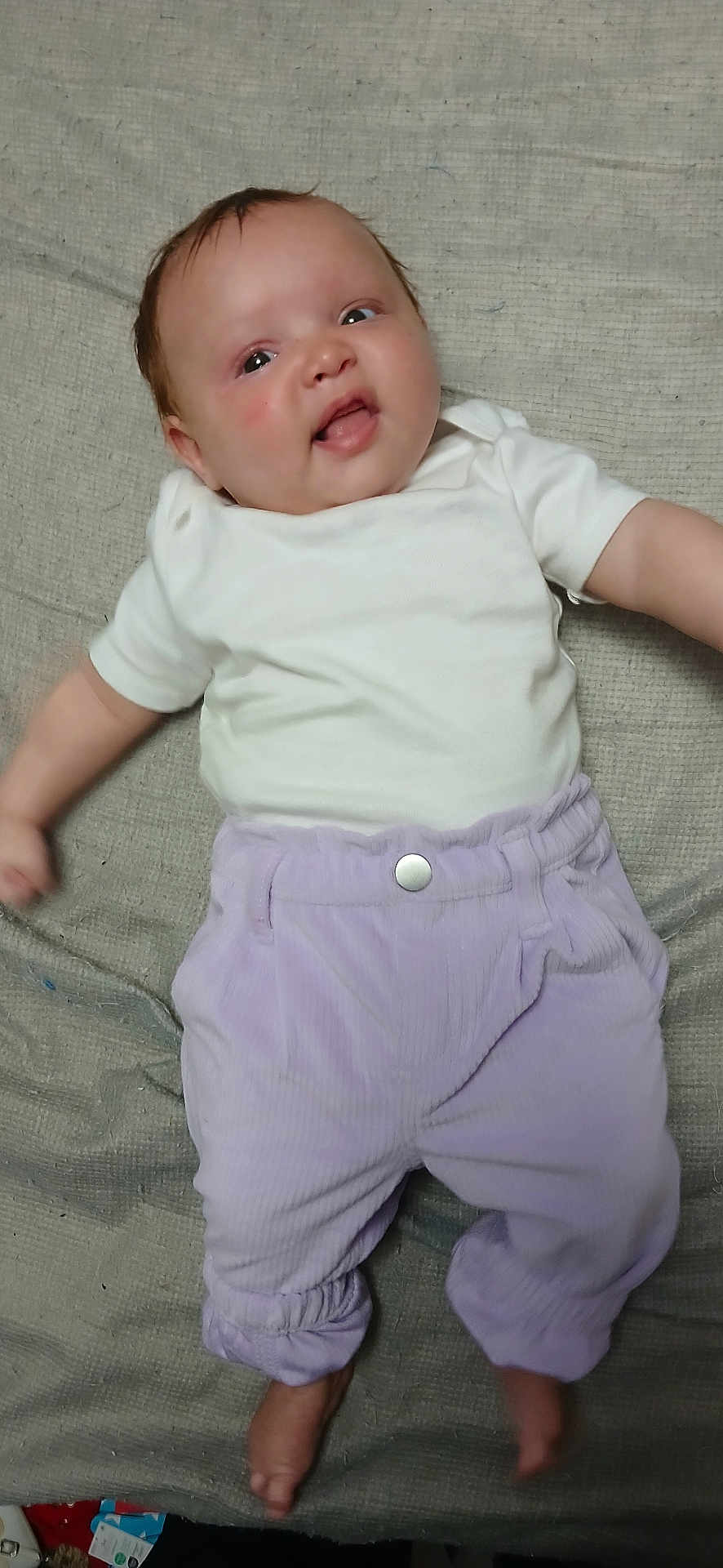 Nala is registered to the contest to win money with this photo: baby, infant, child, white_shirt, purple_pants, bare_feet, lying_down, blanket, textured_background, happy, smiling, tongue_out, face, head, person, clothing, indoors, cute, young, skin