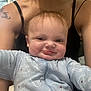 Greyson is registered to the contest to win money with this photo: baby, infant, smiling, blue_onesie, dinosaur_pattern, person, adult, tattoo, tank_top, indoor, window, light, face, expression, cute, child, portrait, young, human, closeup
