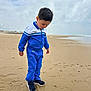 child, boy, beach, sand, tracksuit, blue_clothing, footwear, sneakers, wet_sand, cloudy_sky, sea, water, outdoor, casual_clothing, standing, curious, young_child, nature, footprint, shoreline