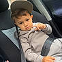 child, car_seat, seatbelt, gray_clothing, cap, car_interior, window, rain_drops, young_child, sitting, safety, casual_wear, thoughtful, indoors, transportation, person, face, hoodie, pants, relaxed