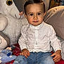 child, toddler, boy, indoor, plush_toy, bed, pillow, jeans, shirt, necklace, cross, socks, toy, curtains, smiling, sitting, comfortable, cute, portrait, home