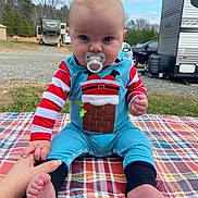 Montgomery is registered to the contest to win money with this photo: baby, infant, child, sitting, pacifier, christmas_outfit, festive, onesie, striped_sleeves, smiling, portrait, outdoor, picnic_blanket, campground, trailer, rv, vehicles, trees, sky, bare_feet
