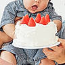 baby, child, birthday, cake, strawberries, dessert, sitting, highchair, portrait, indoors, smiling, sleepy, open_mouth, striped_clothing, bare_feet, adult_hands, celebration, white_background, food, family