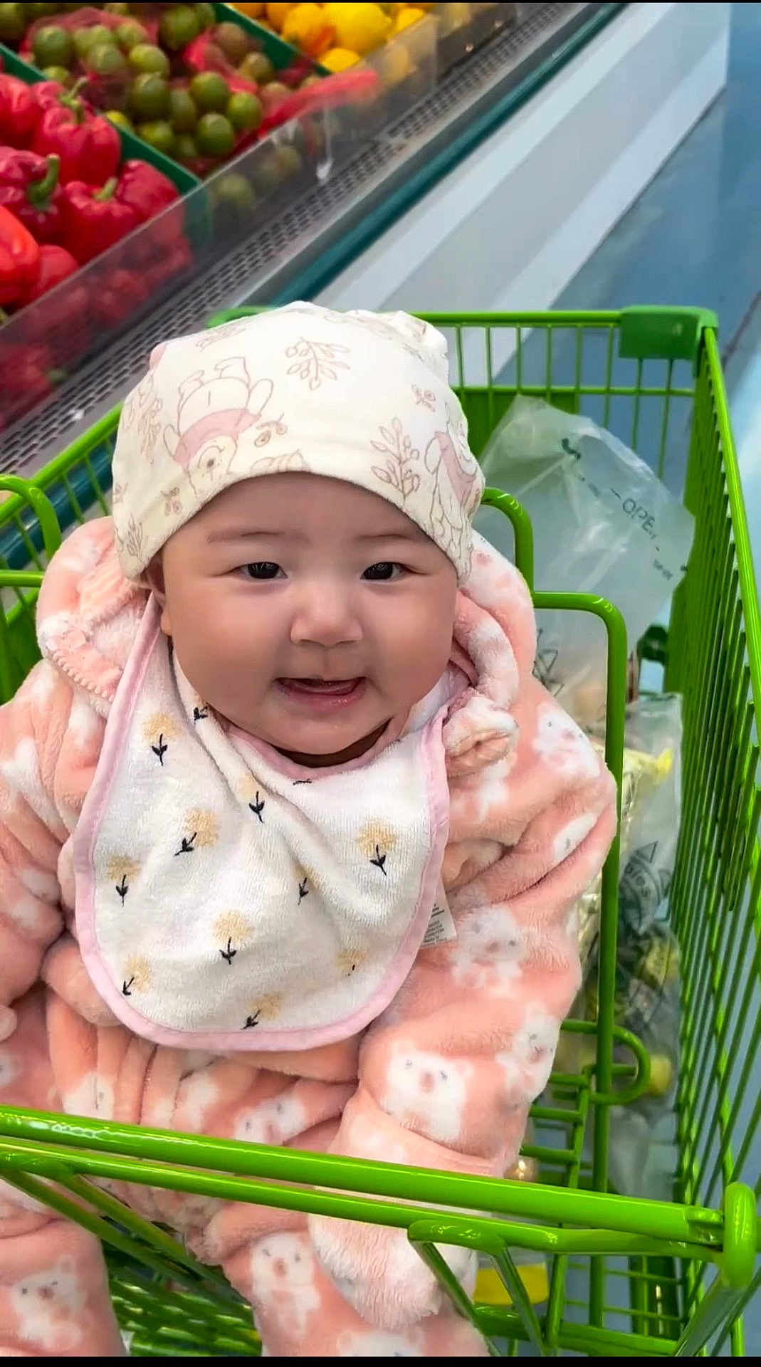 Naomi joined the competition — help win amazing prizes! baby, child, shopping_cart, grocery_store, produce, red_peppers, green_peppers, bib, hat, pink_onesie, smiling, face, cute, indoor, person, clothing, food, shopping, happy, infant