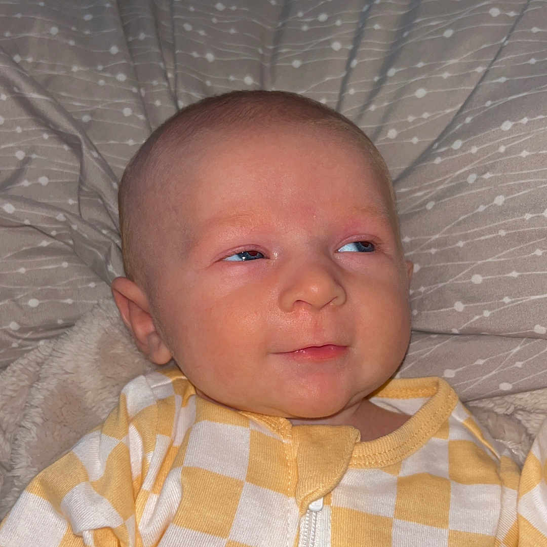 Winston is registered to the contest to win money with this photo: baby, child, infant, onesie, yellow, white, checkered, pillow, blanket, face, head, skin, eyes, expression, lying_down, indoors, portrait, cute, smile, baby_clothes