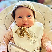 Athena is registered to the contest to win money with this photo: baby, infant, child, hat, bib, clothing, face, person, portrait, cute, wide_eyes, indoor, seat, fabric, soft_tones, newborn, hand, cozy, smile, young