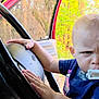 toddler, child, baby, pacifier, steering_wheel, car_interior, hand, face, expression, serious, blue_shirt, seat, window, door, outdoor, nature, trees, daylight, person, vehicle