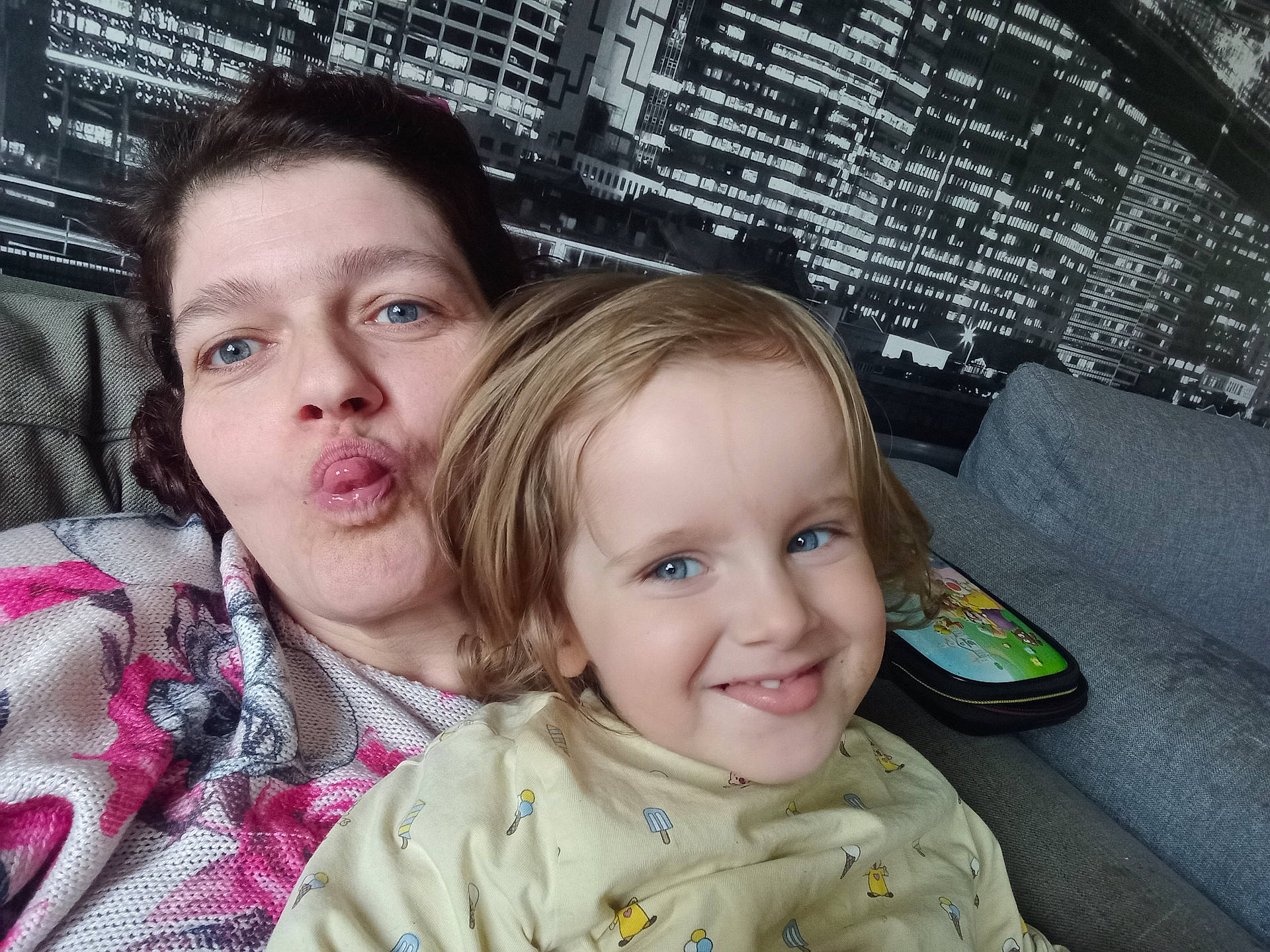 Jasmine a rejoint le concours — aidez-le/la à gagner de superbes lots ! adult, blond_hair, blue_eyes, casual, child, cityscape, couch, family, funny_face, grey_couch, happy, home, indoor, light, mural, patterned_clothing, portrait, selfie, smile, tongue_out