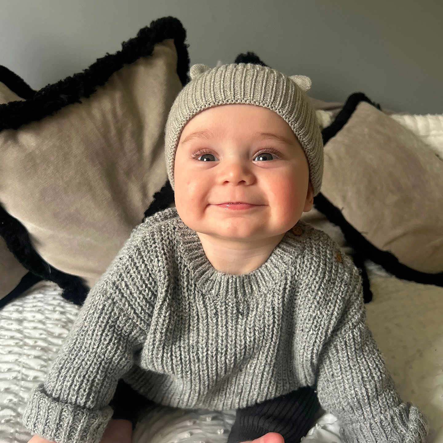 Reggie joined the competition — help win amazing prizes! baby, blanket, child, cozy, cushion, cute, face, foot, grey_sweater, hand, happy, home, indoors, infant, knitted_hat, portrait, sitting, smiling, soft_lighting, warm_clothing