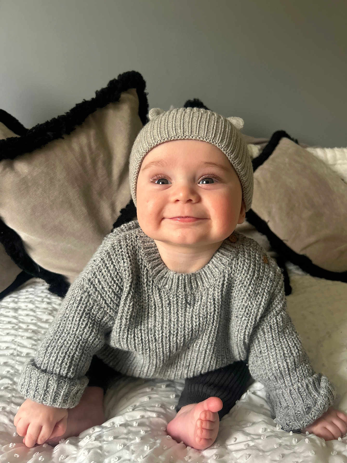 Reggie joined the competition — help win amazing prizes! baby, child, smiling, knitted_hat, grey_sweater, blanket, cushion, cozy, indoors, cute, happy, portrait, infant, sitting, soft_lighting, home, warm_clothing, foot, hand, face