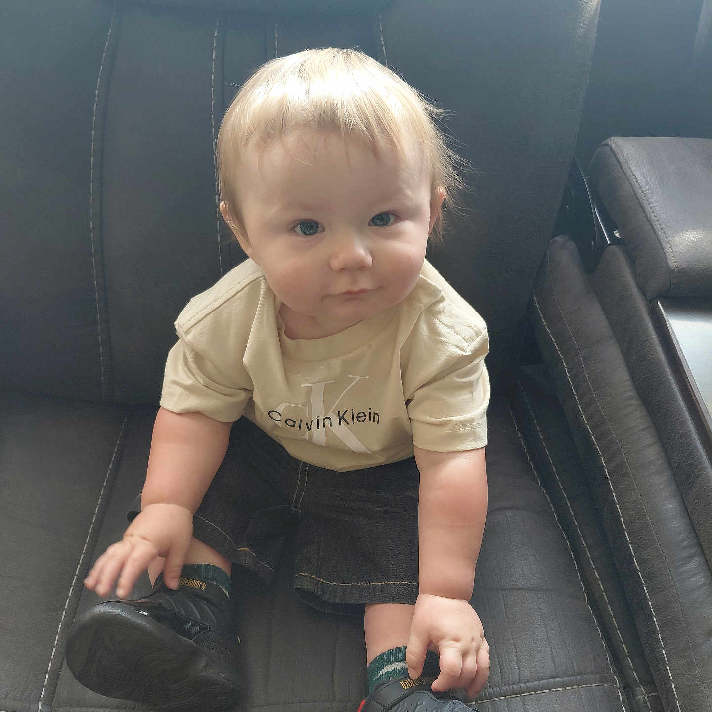 Isaac is registered to the contest to win money with this photo: baby, child, toddler, portrait, sitting, car, car_seat, vehicle, blond_hair, blue_eyes, serious_expression, face, clothing, shirt, logo, shorts, sneakers, shoes, indoor, photography