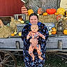 Randy is registered to the contest to win money with this photo: person, baby, child, pumpkins, hay_bales, wagon, tree, autumn, fall, outdoor, smiling, clothing, nature, seasonal, festive, orange, green, red_barn, grass, lantern