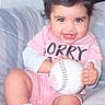 baby, baseball, child, couch, cozy, curly_hair, cute, happy, holding_object, indoor, infant, person, pink_clothing, playful, portrait, seated, smiling, socks, soft_lighting, young_child