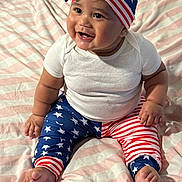 Dior is registered to the contest to win money with this photo: baby, infant, smiling, sitting, patriotic, stars_and_stripes, clothing, hat, leggings, white_shirt, bed, blanket, cute, happy, chubby, person, indoors, face, foot, hand