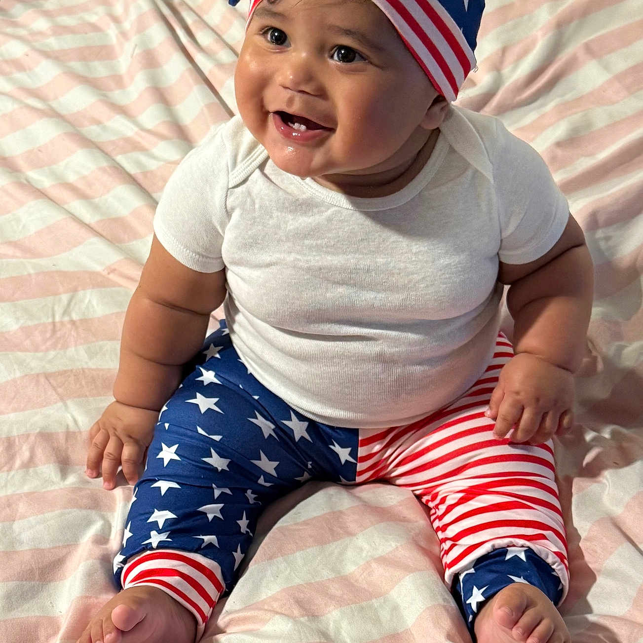 Dior is registered to the contest to win money with this photo: baby, infant, smiling, sitting, patriotic, stars_and_stripes, clothing, hat, leggings, white_shirt, bed, blanket, cute, happy, chubby, person, indoors, face, foot, hand