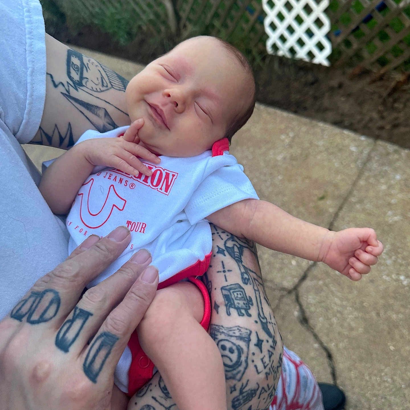 Meadow is registered to the contest to win money with this photo: arm, baby, clothing, cute, fence, garden, hand, holding, infant, newborn, outdoor, peaceful, person, red_print, relaxing, skin, smile, sock, tattoos, white_shirt