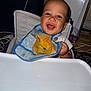 baby, smiling, high_chair, bib, lion_image, indoor, happy, child, face, person, furniture, appliance, refrigerator, cables, seat, table, clothing, infant, cute, portrait