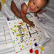 Julien is registered to the contest to win money with this photo: baby, child, paint, canvas, colorful, smiling, face_paint, art, messy, white_sheet, palette, paintbrush, sitting, happy, infant, hand_paint, creative, indoors, person, playful