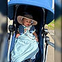 baby, stroller, hat, blanket, pacifier, outdoor, child, smiling, sunlight, clothing, blue, person, baby_boy, accessory, happy, infant, pavement, grass, daylight, comfort
