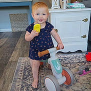 Eadie is registered to the contest to win money with this photo: barefoot, blue_table, cabinet, carpet, child, clothing, flooring, furniture, happy, home_interior, indoor, person, play, red_hair, ride_on_toy, rug, smiling, toddler, toy_phone, wood_floor