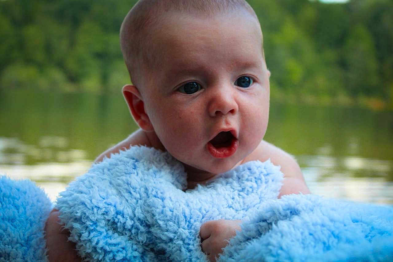 Noah is registered to the contest to win money with this photo: baby, blue_blanket, face, infant, outdoor, water, nature, trees, expression, skin, eyes, mouth, hand, portrait, candid, child, young, soft_texture, closeup, serene