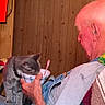 cat, gray_cat, person, elderly_man, indoors, wood_paneling, couch, blanket, bowl, spoon, sitting, pet, human_animal_interaction, portrait, home, cozy, framed_picture, casual, vertical_photo, funny