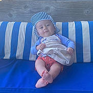 Léandre a rejoint le concours — aidez-le/la à gagner de superbes lots ! baby, child, cap, striped_clothing, shorts, cushion, blue, white, striped_pattern, bag, wood, bench, outdoor, smile, feet, person, sitting, relaxed, cute, portrait