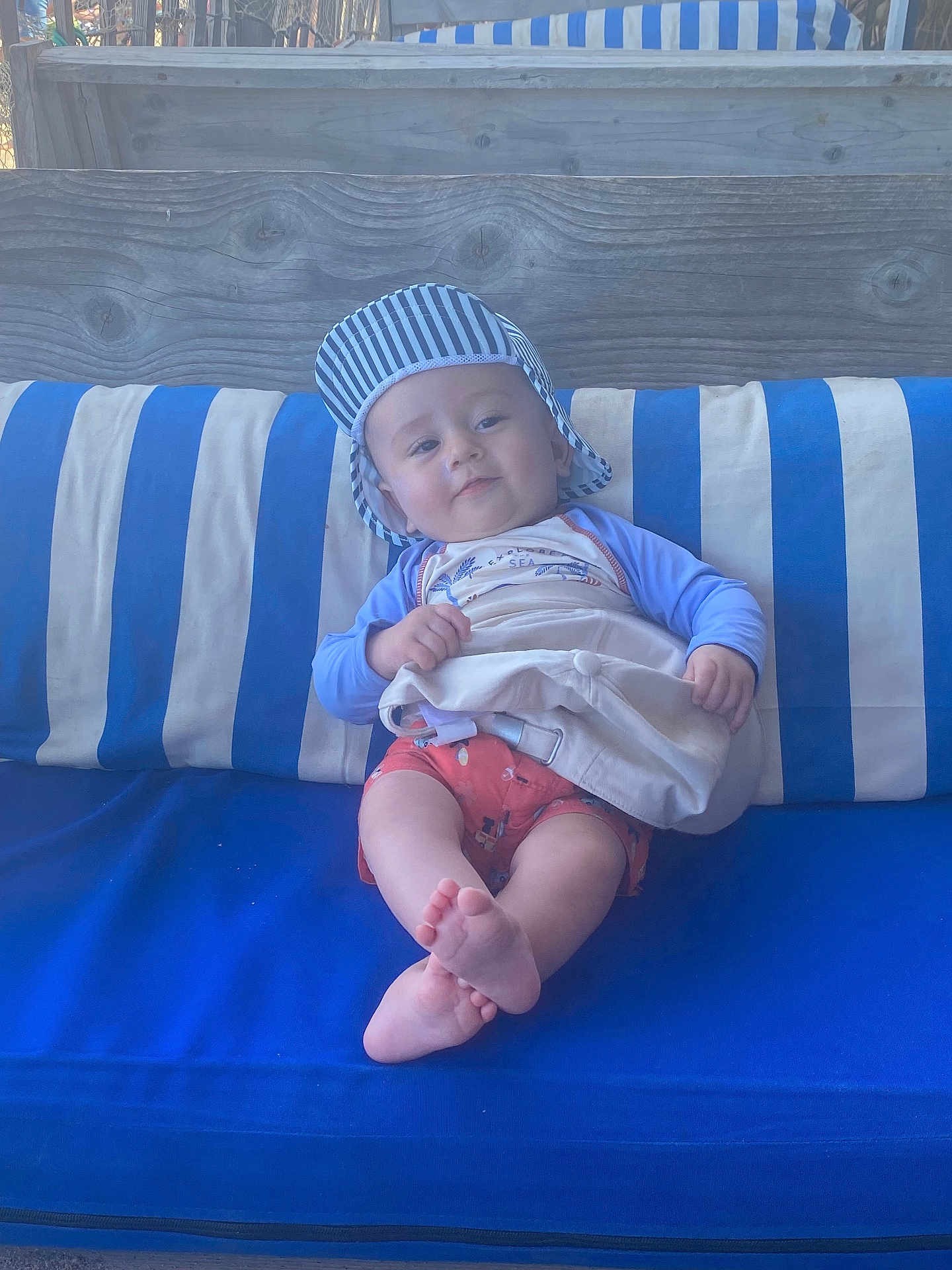 Léandre a rejoint le concours — aidez-le/la à gagner de superbes lots ! baby, child, cap, striped_clothing, shorts, cushion, blue, white, striped_pattern, bag, wood, bench, outdoor, smile, feet, person, sitting, relaxed, cute, portrait