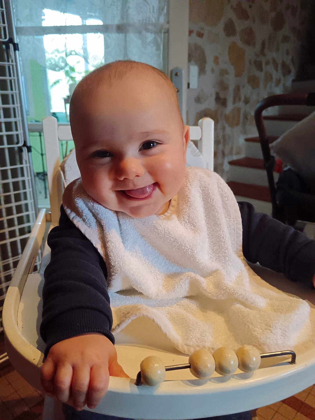 Malo a rejoint le concours — aidez-le/la à gagner de superbes lots ! baby, child, smiling, high_chair, bib, indoor, hand, face, tongue, happy, young, furniture, stone_wall, window, natural_light, person, cute, playful, infant, grip