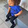 child, curly_hair, blue_shirt, black_pants, bench, tile_floor, indoor, casual_clothing, seated, thoughtful, person, young, modern_interior, footwear, looking_away, hairstyle, contemplative, monochrome_floor, relaxed_pose, daylight