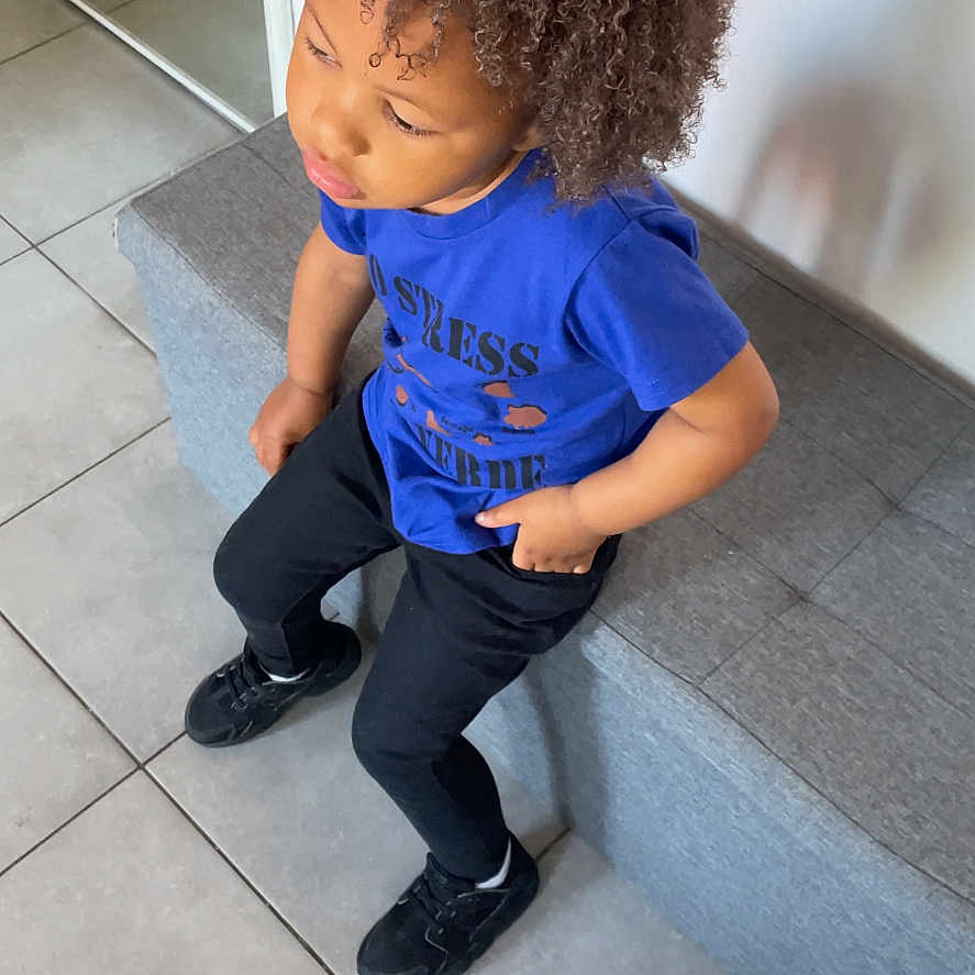 Deyvon participe au concours pour gagner de l'argent avec cette photo : child, curly_hair, blue_shirt, black_pants, bench, tile_floor, indoor, casual_clothing, seated, thoughtful, person, young, modern_interior, footwear, looking_away, hairstyle, contemplative, monochrome_floor, relaxed_pose, daylight