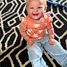 baby, child, smiling, happy, sitting, jeans, orange_top, patterned_rug, indoor, cute, blonde_hair, blue_eyes, barefoot, portrait, casual_clothing, young_child, infant, person, home, floor