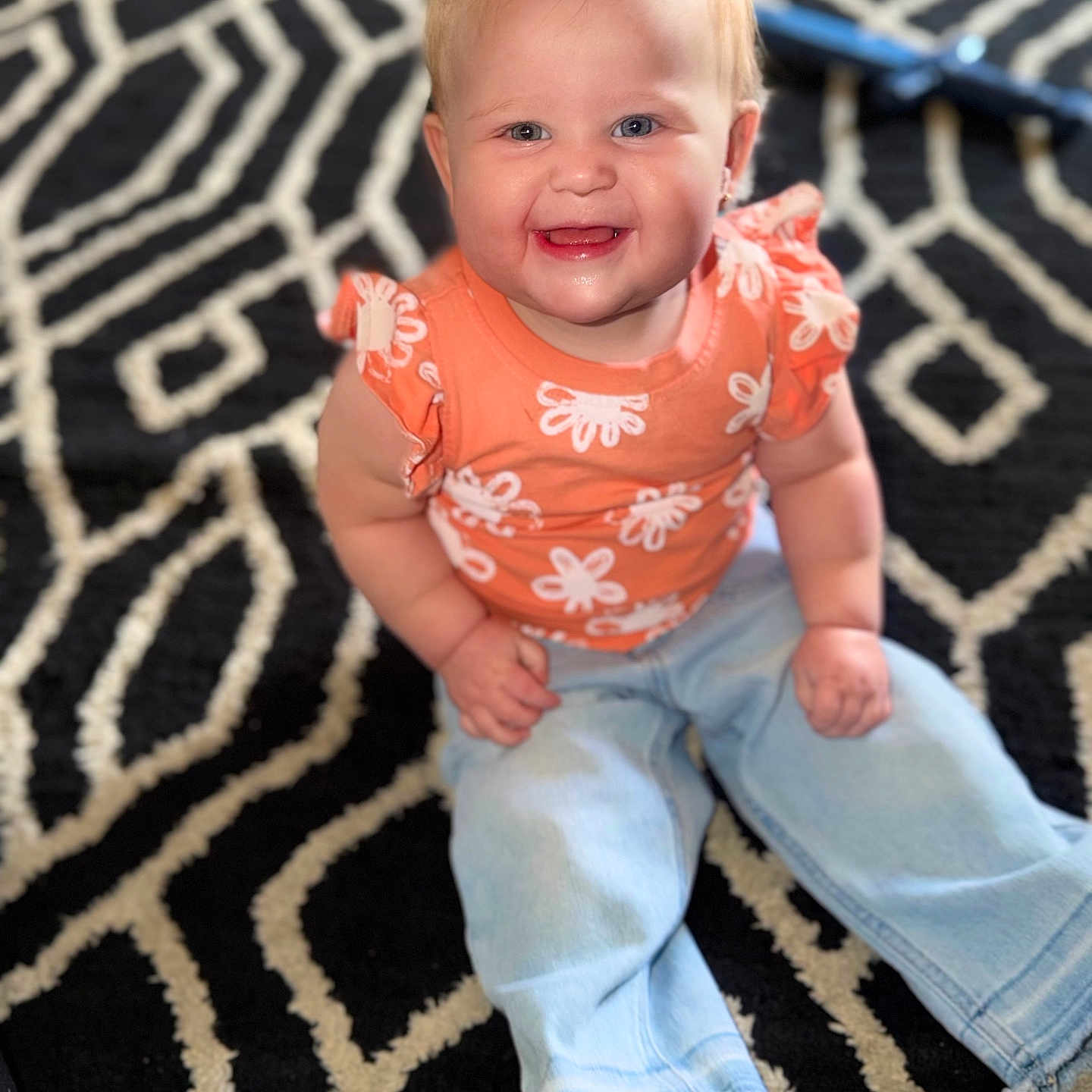 Everleigh joined the competition — help win amazing prizes! baby, child, smiling, happy, sitting, jeans, orange_top, patterned_rug, indoor, cute, blonde_hair, blue_eyes, barefoot, portrait, casual_clothing, young_child, infant, person, home, floor