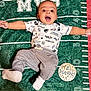 Kai is registered to the contest to win money with this photo: age_marker, arms_outstretched, baby, child, clothing, cute, face, football_field_design, green_blanket, happy, indoor, infant, lying_down, pants, person, portrait, shirt, smiling, socks, wooden_sign