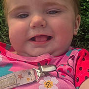 Rayven is registered to the contest to win money with this photo: baby, child, close_up, smiling, pink_clothing, flower_pattern, ladybug_pattern, grass, outdoor, face, cheeks, tongue, pacifier_clip, young_child, happy, cute, infant, head, skin, person