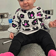 Dakota is registered to the contest to win money with this photo: baby, black_pants, child, clothing, cute, electrical_outlet, exit_sign, expression, face, hair, indoor, metal_table, person, pigtails, pink_socks, playful, sitting, smiling, sweater, water_bottle