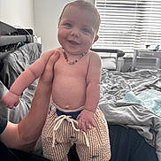 Joseph is registered to the contest to win money with this photo: baby, child, smiling, shirtless, shorts, necklace, hand, bedroom, bed, blanket, window, blinds, person, indoor, happy, cute, infant, skin, foot, adult_hand