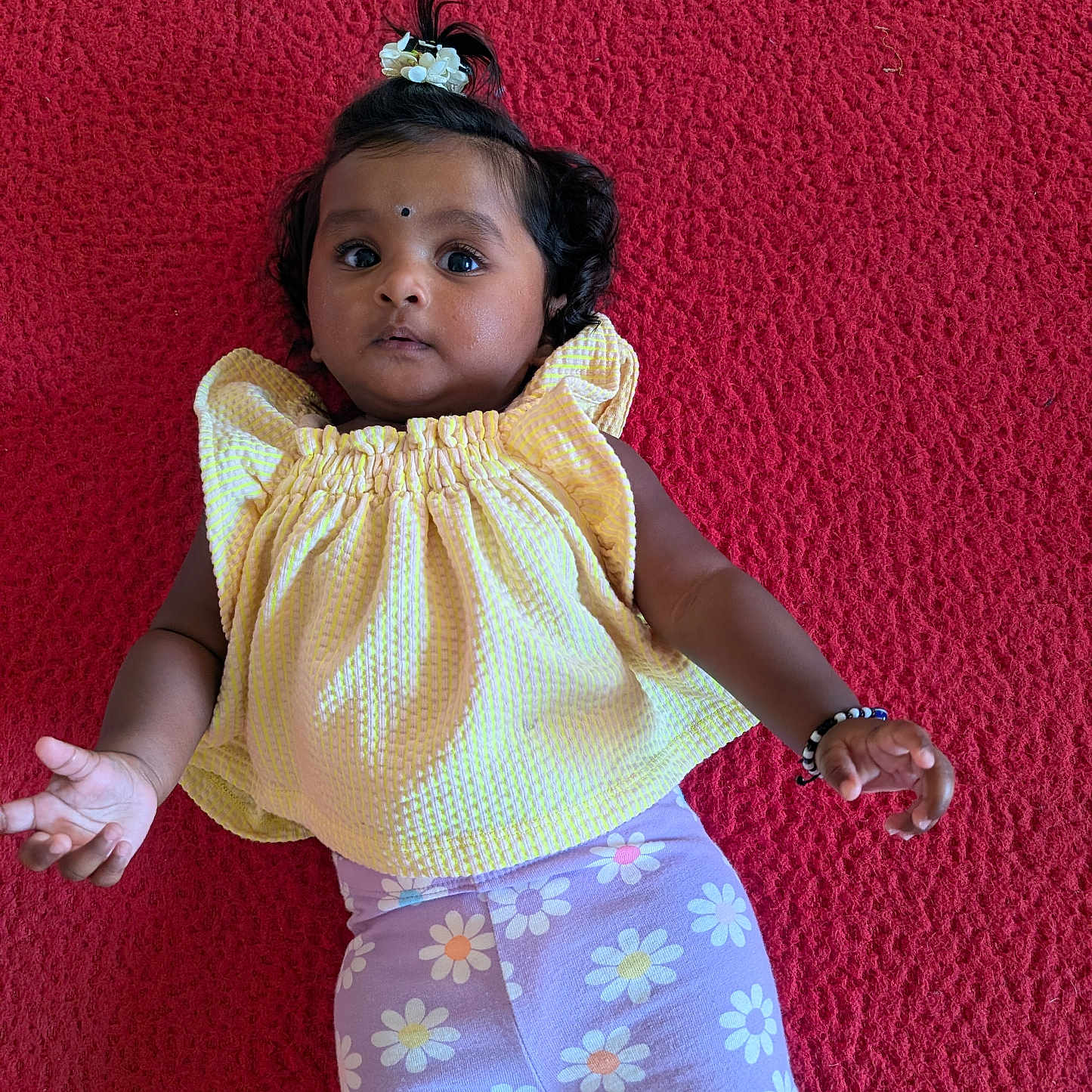 Sai Ishanvi joined the competition — help win amazing prizes! baby, casual, child, colorful_clothing, curious_expression, cute, dark_hair, face, floral_shorts, hair_accessory, indoors, infant, lying_down, person, portrait, red_background, skin, small_hands, yellow_top, young_child
